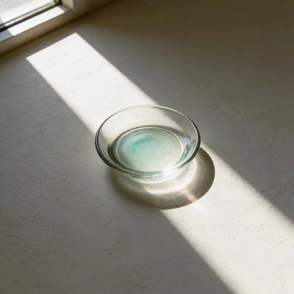 A shallow glass dish with a few millimetres of pale blue-green liquid in it, on a warm off-white surface.
